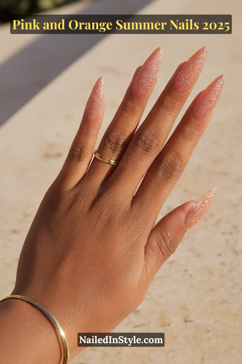 Pink and Orange Summer Nails 2026: Sunset Tips, Endless Vibes 10 Hand with long almond-shaped nails featuring a glitter gradient of pink and orange starting at the cuticle, posed palm-down on a sunlit stone surface with warm-toned skin.