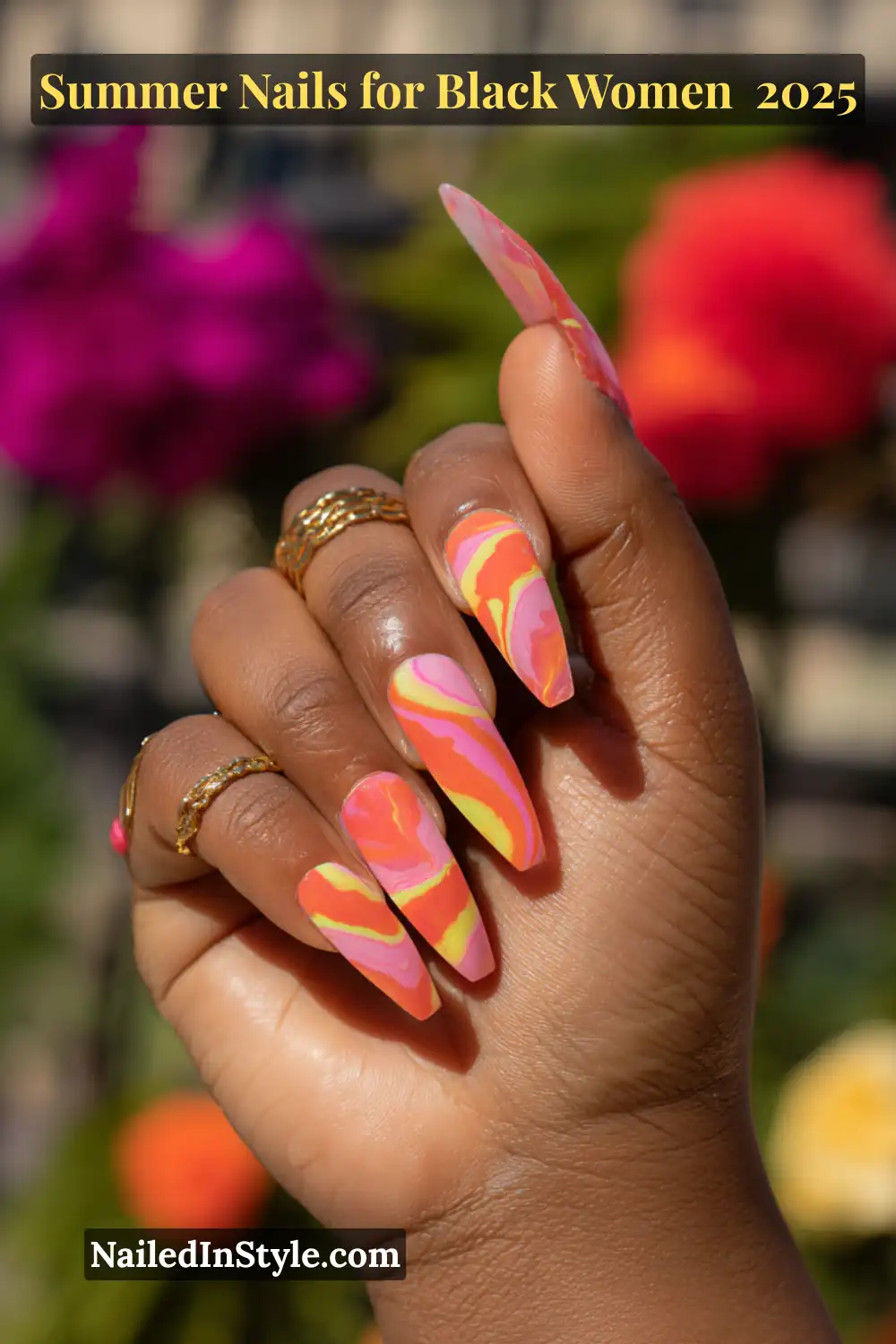 Black woman's hand with long stiletto nails in neon pink, orange, and yellow swirls, set against colorful flowers and gold rings.