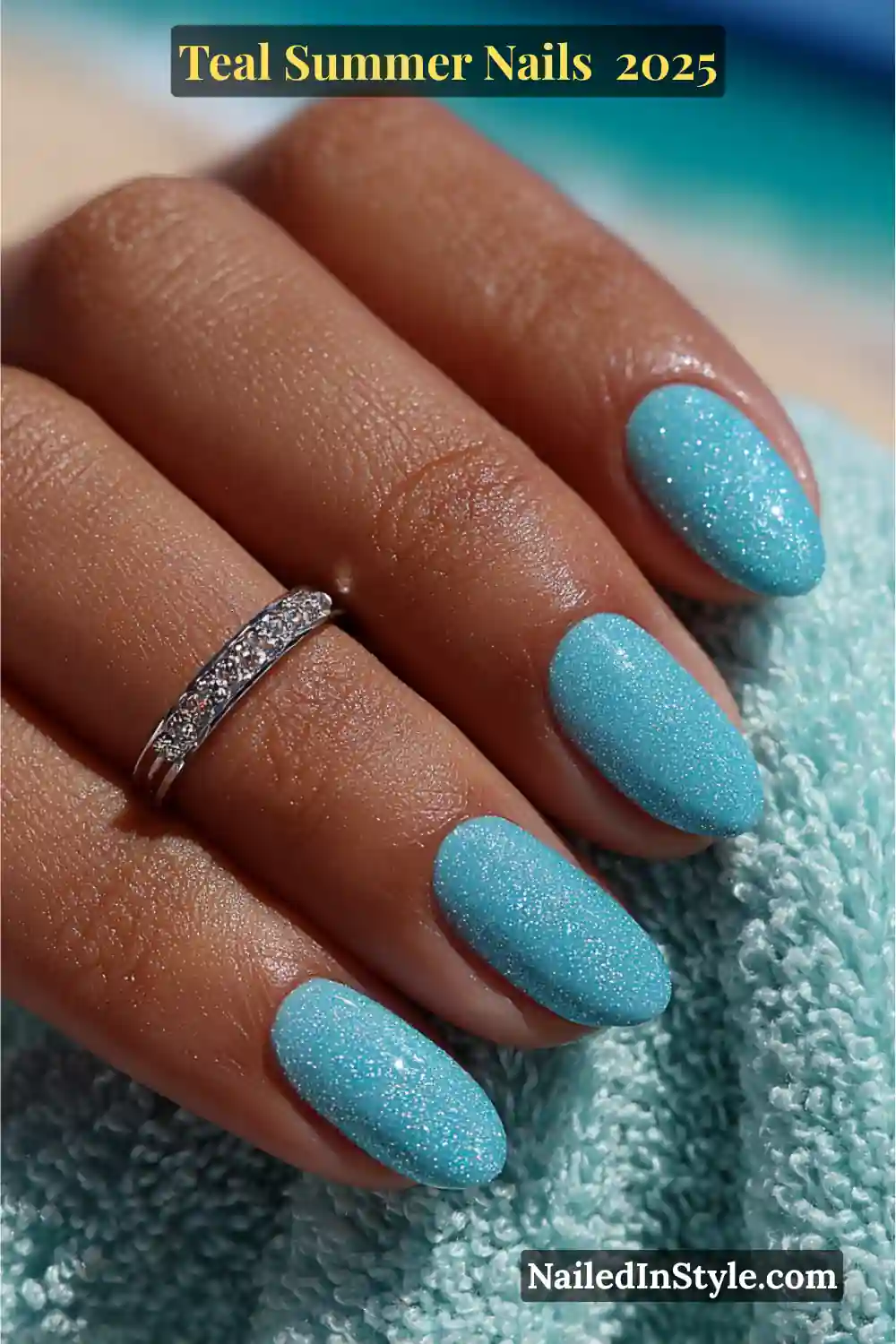 Close-up of soft oval-shaped nails in shimmering teal polish, styled on a medium-deep hand with a silver ring and turquoise towel backdrop
