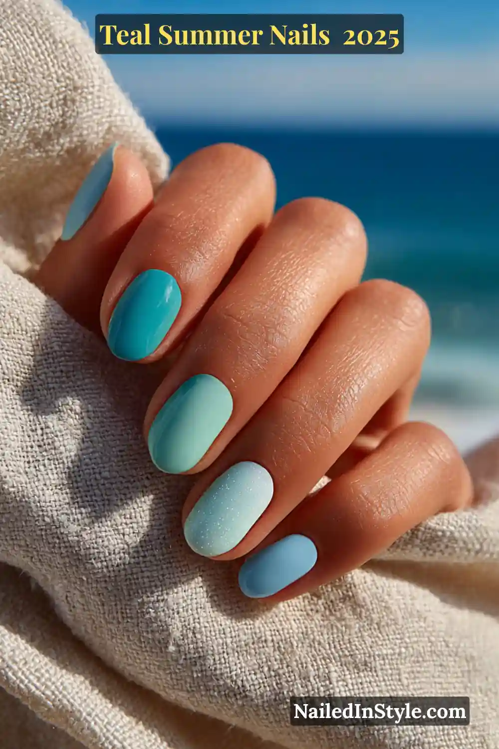 Natural female hand with short oval nails in a teal to baby blue gradient polish, resting on linen fabric with ocean waves in the background