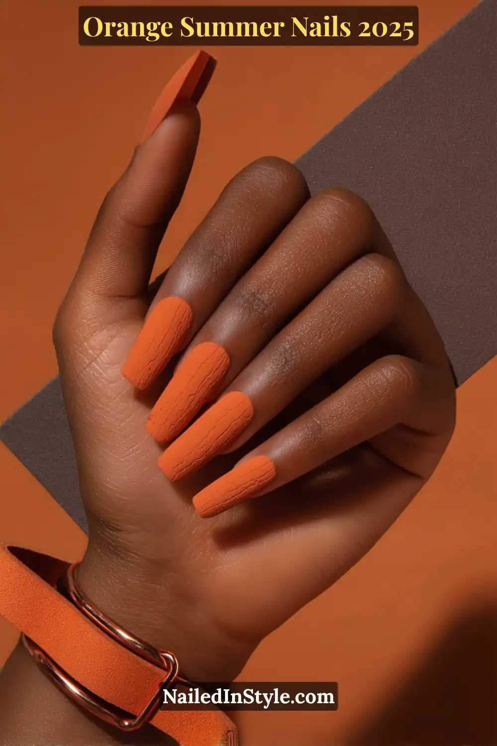 Close-up of hand with long square matte orange gel nails featuring a subtle embossed texture, styled with an orange strap bracelet and warm background.