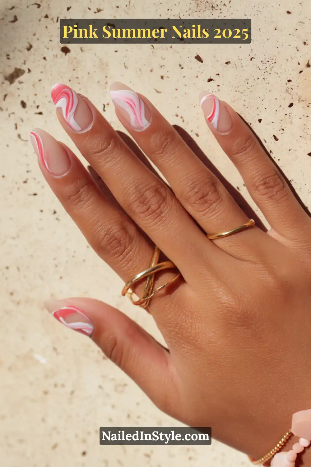 Close-up of a hand with almond-shaped nails featuring pink and white abstract swirl art on a soft nude matte base, accented with gold and beaded bracelets.