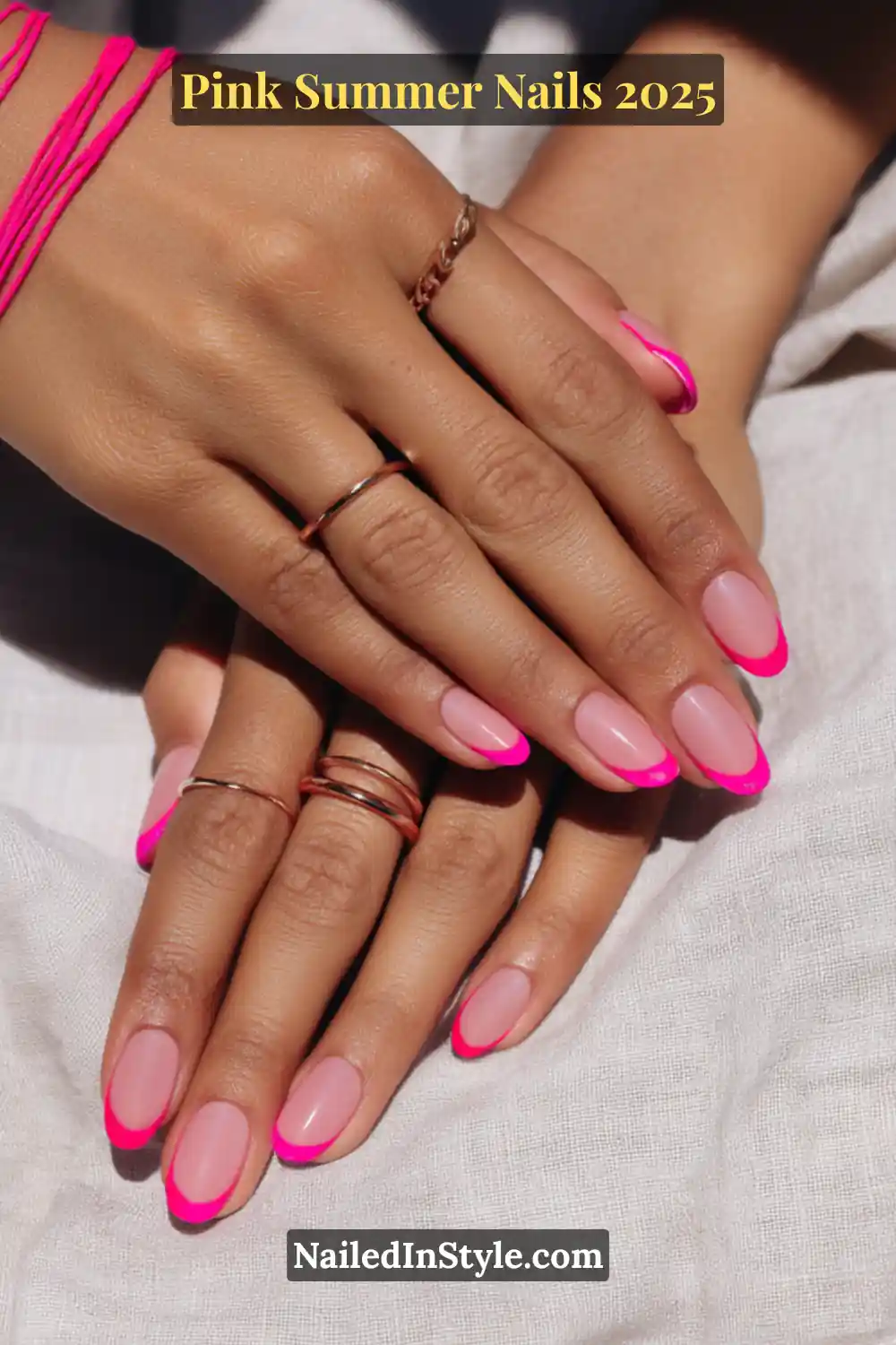 Hands with oval nails featuring vivid pink French tips and a nude base, styled with rose gold rings and pink string bracelets.