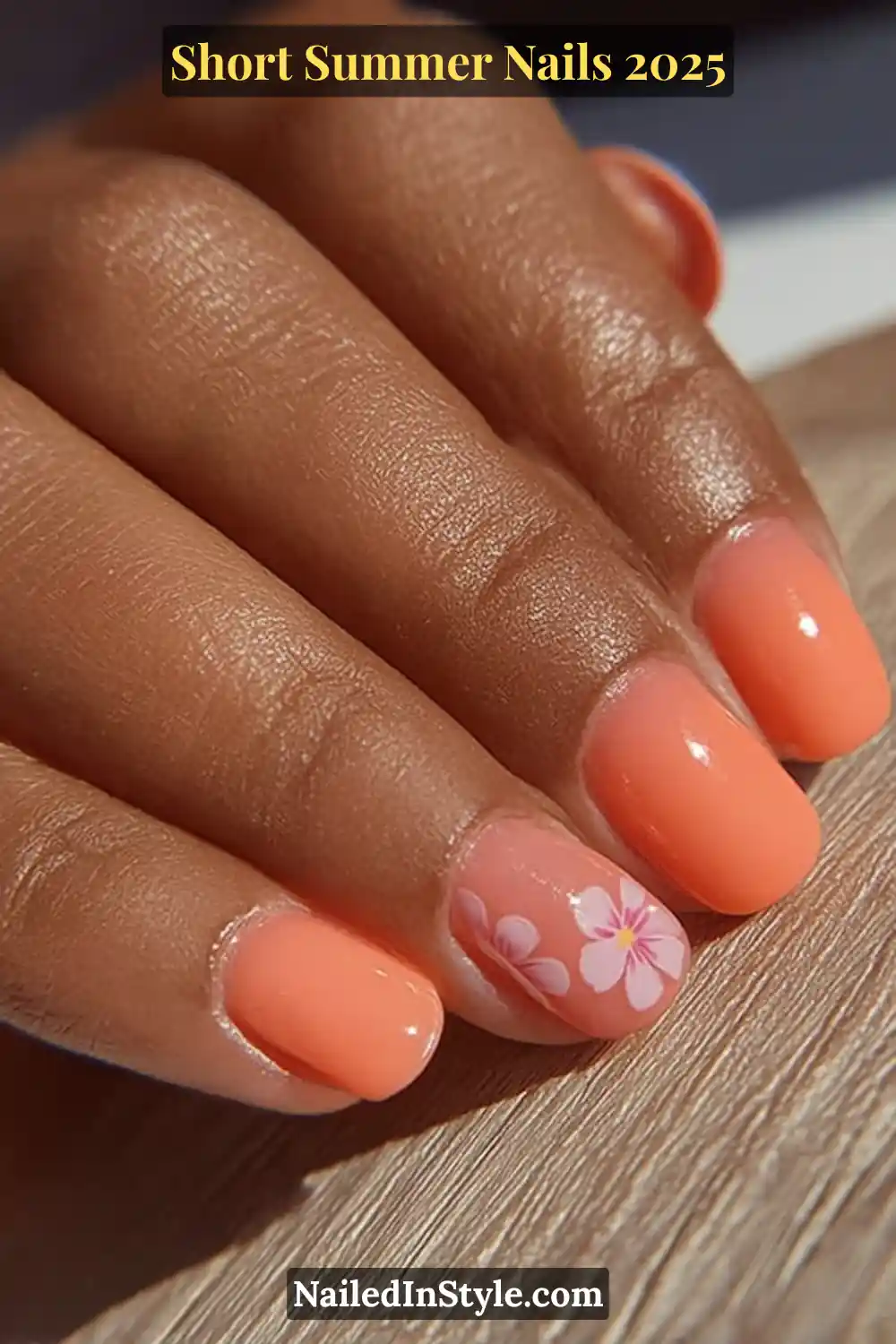 Glossy peach-toned nails on a deep skin hand, featuring a floral accent nail with delicate pink hibiscus-inspired blossoms, photographed in natural sunlight on a wooden surface.