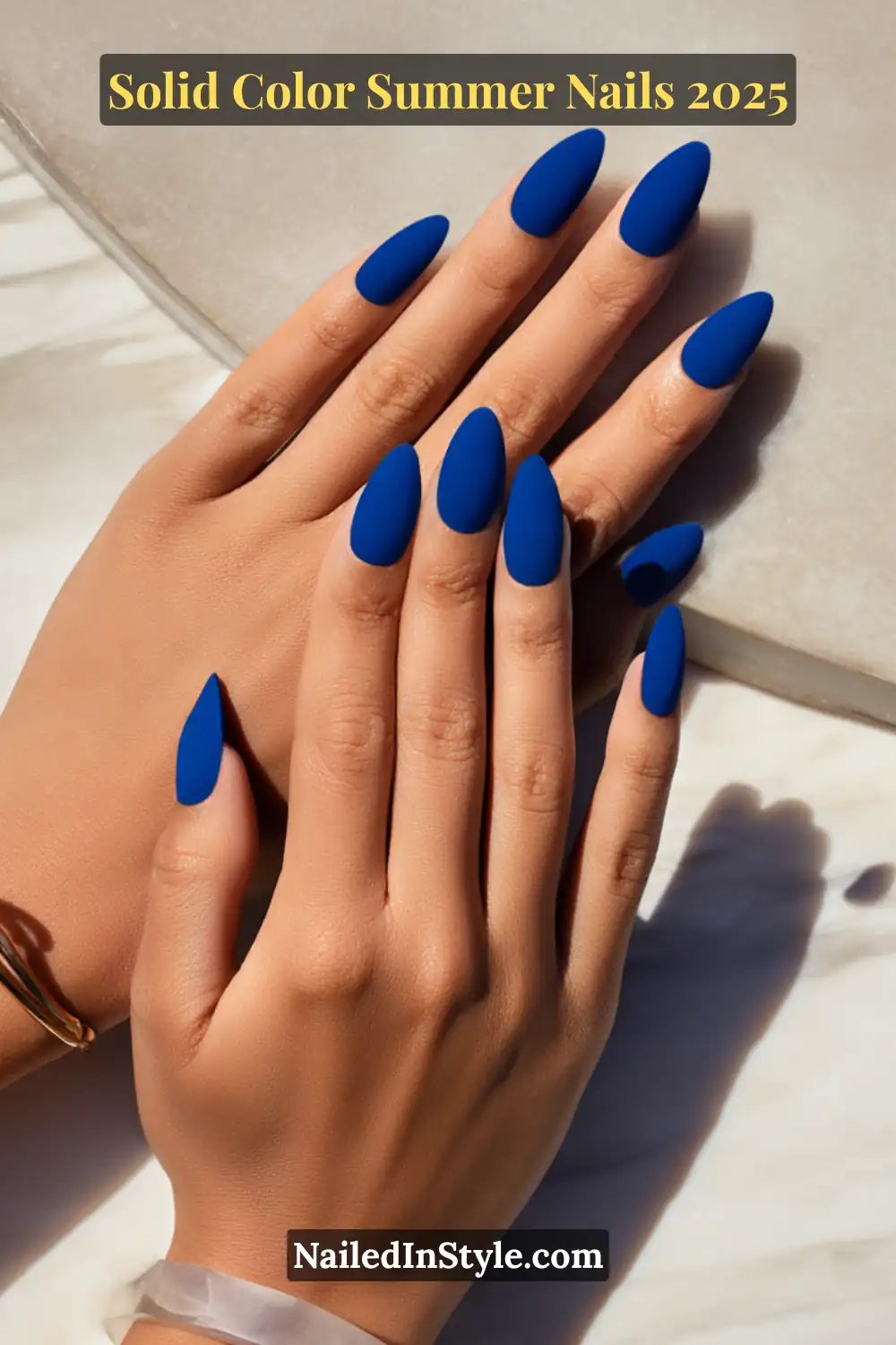 Hands with almond-shaped matte cobalt blue nails resting on a stone surface in soft sunlight.