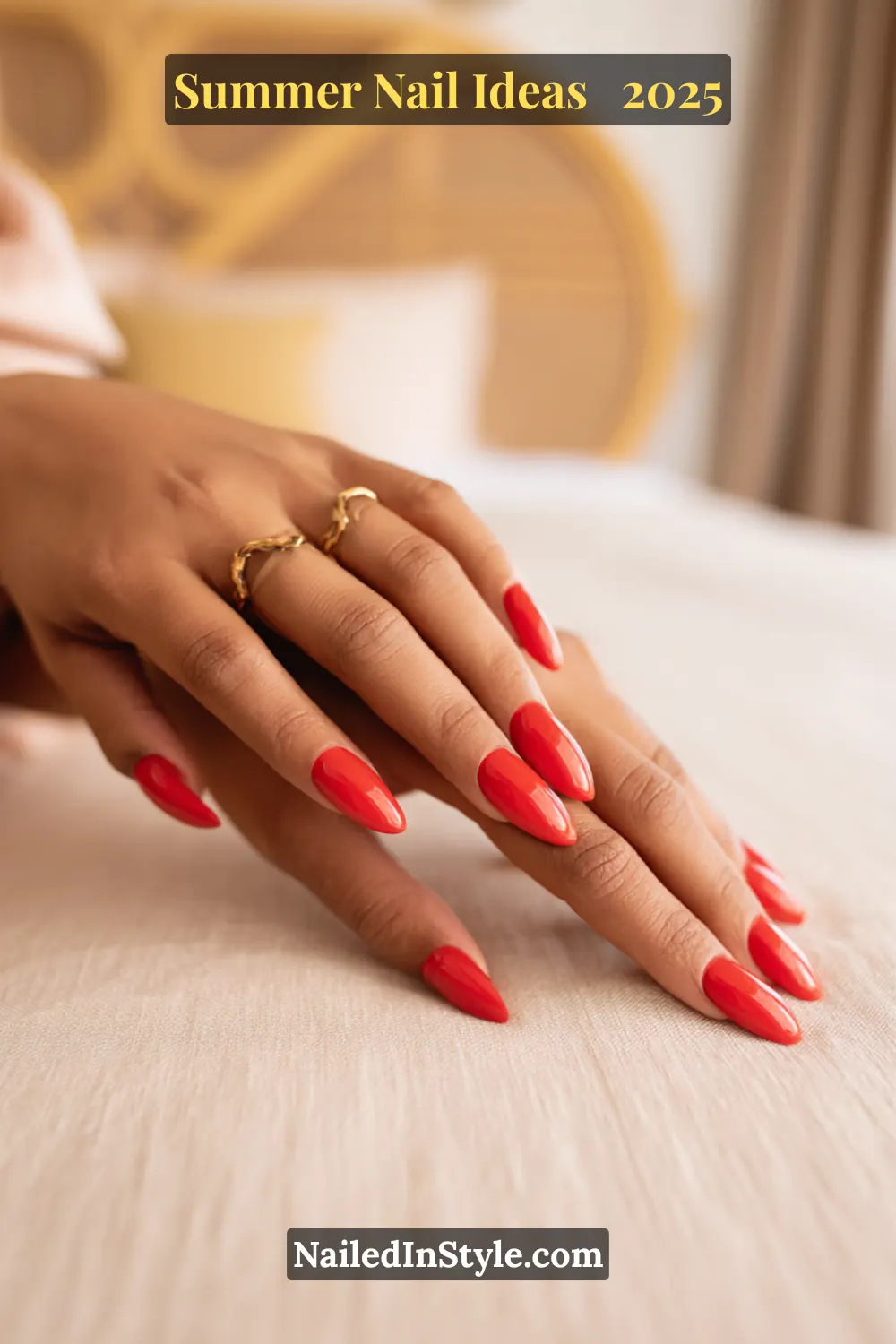 Close-up of warm-toned hands with sharp stiletto nails painted in a high-shine coral red BIAB (Builder in a Bottle) gel polish, posed on a neutral fabric surface with soft golden decor in the background.