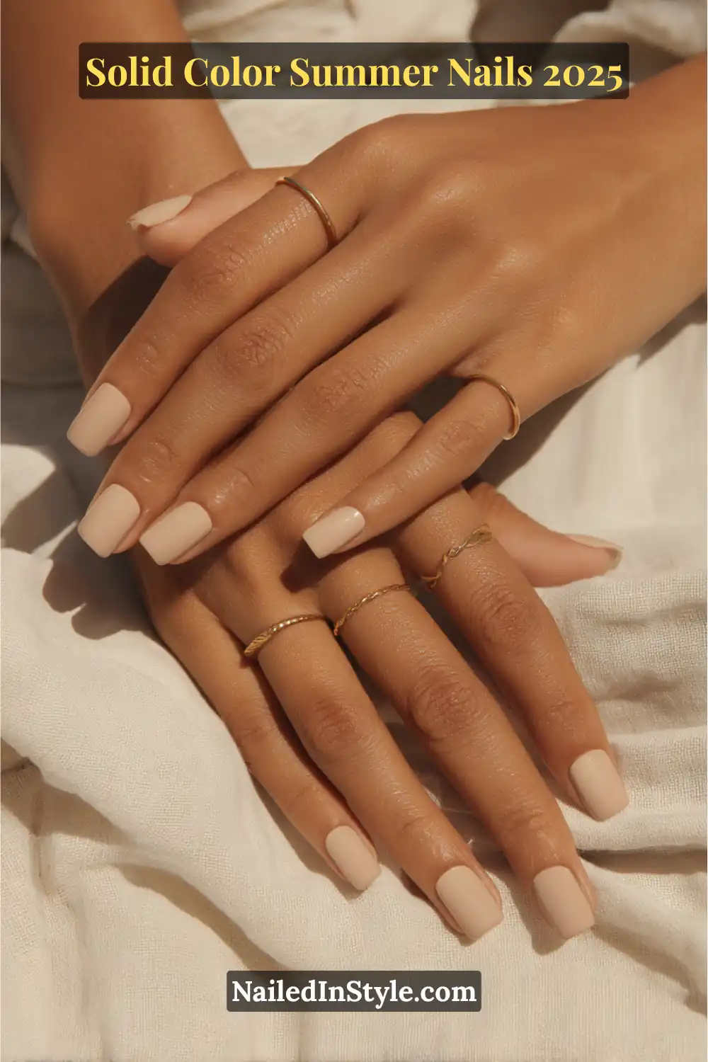 Two hands with short square nails painted in a matte warm sand nude polish, styled with gold rings on sunlit fabric.