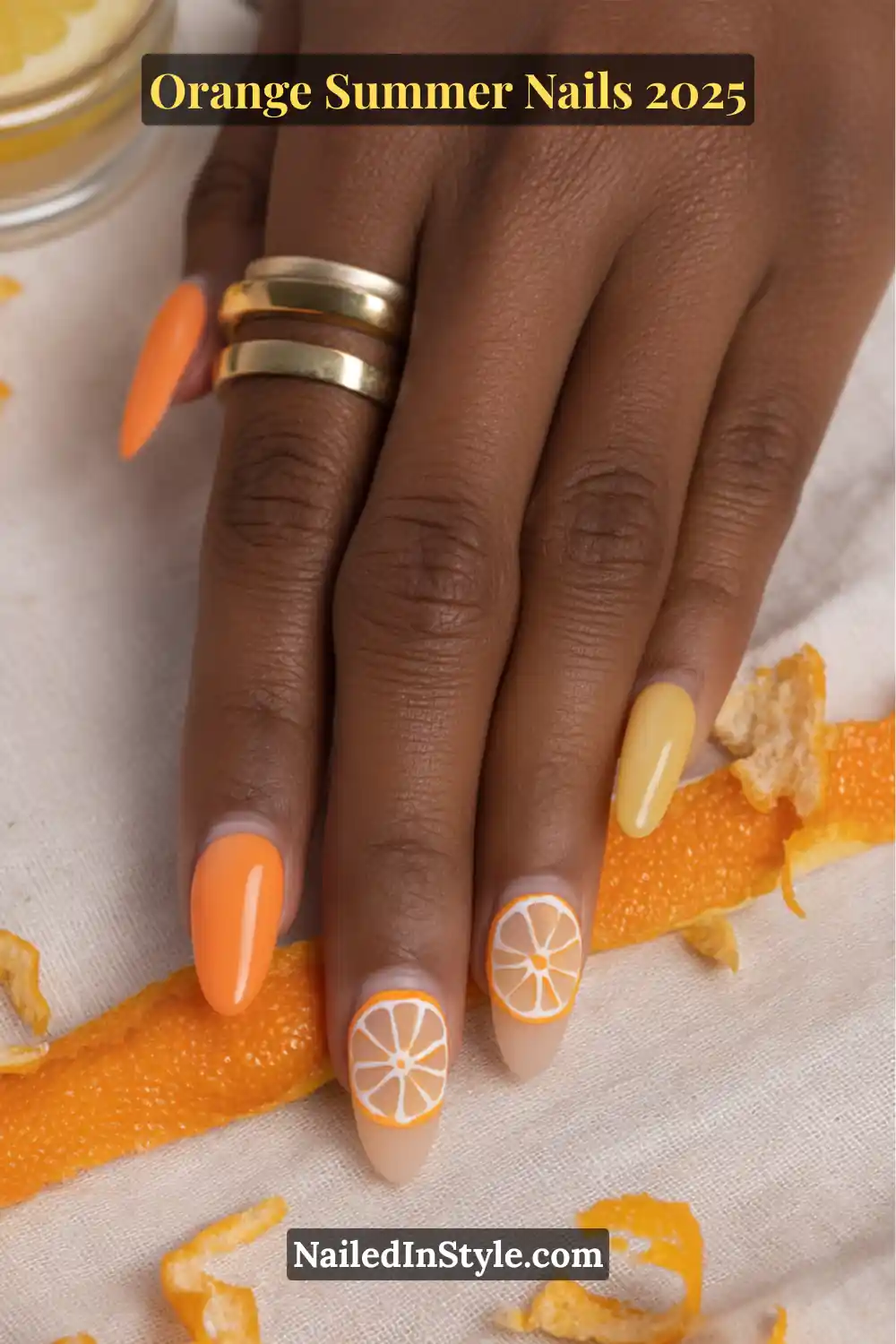 Close-up of a hand with orange and yellow summer nails, featuring two accent nails with citrus slice designs in white and orange.