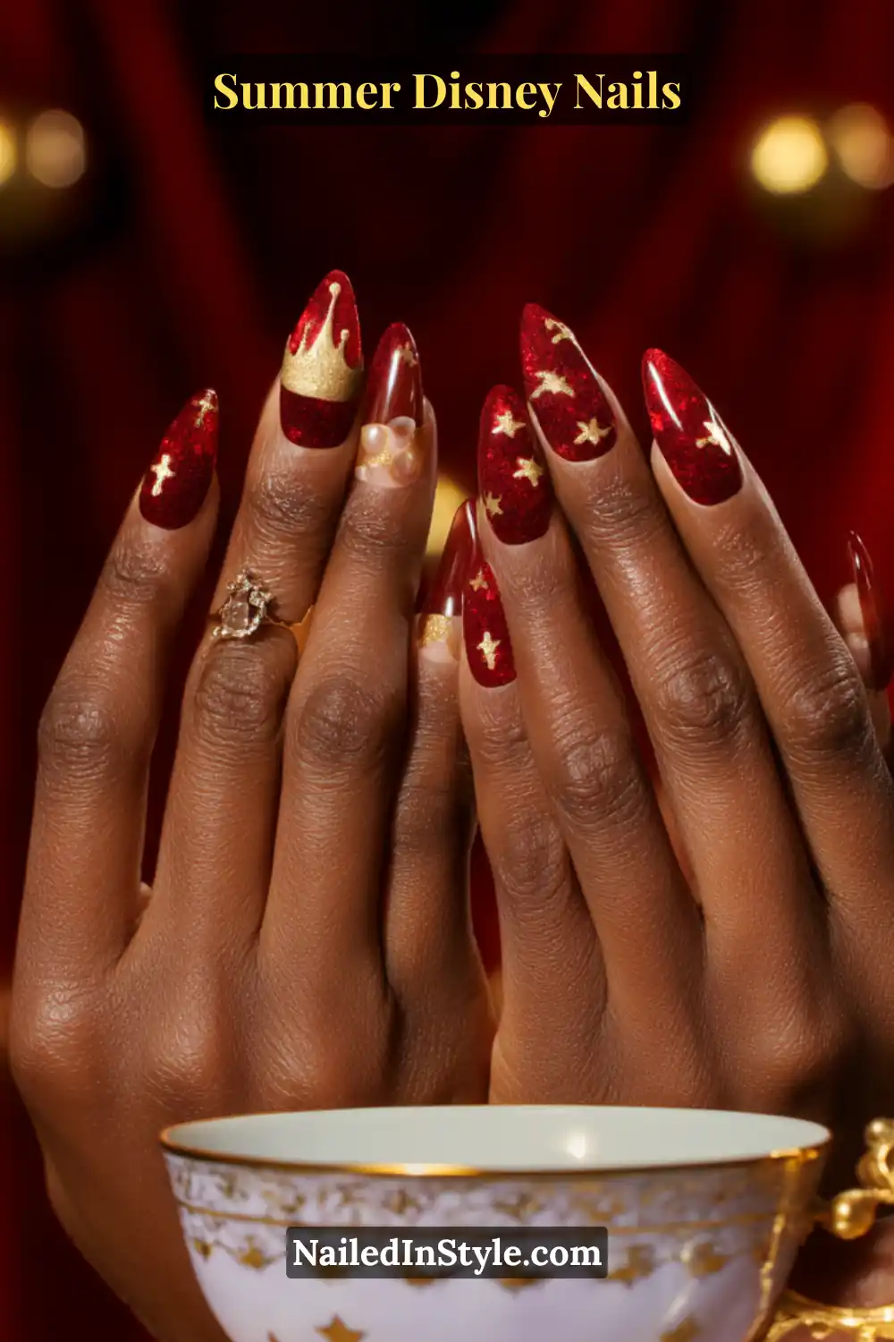 Long almond-shaped nails in deep red with gold crowns, stars, and Mickey accents, glowing against deep skin.