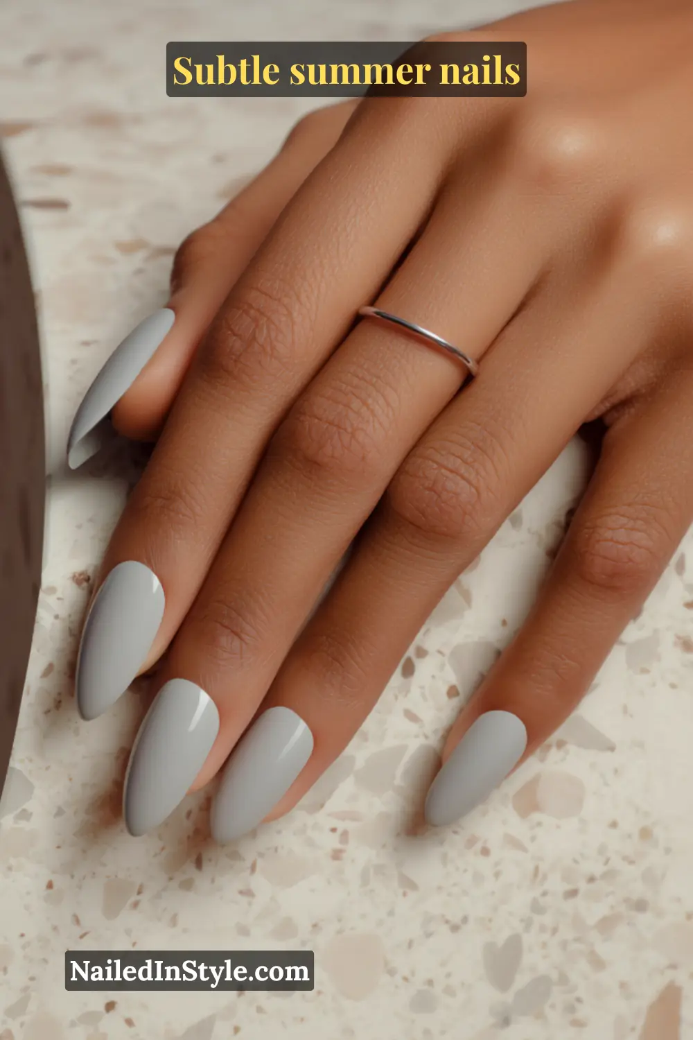 Glossy almond-shaped nails painted in a soft dove gray shade, resting gracefully on a terrazzo surface.