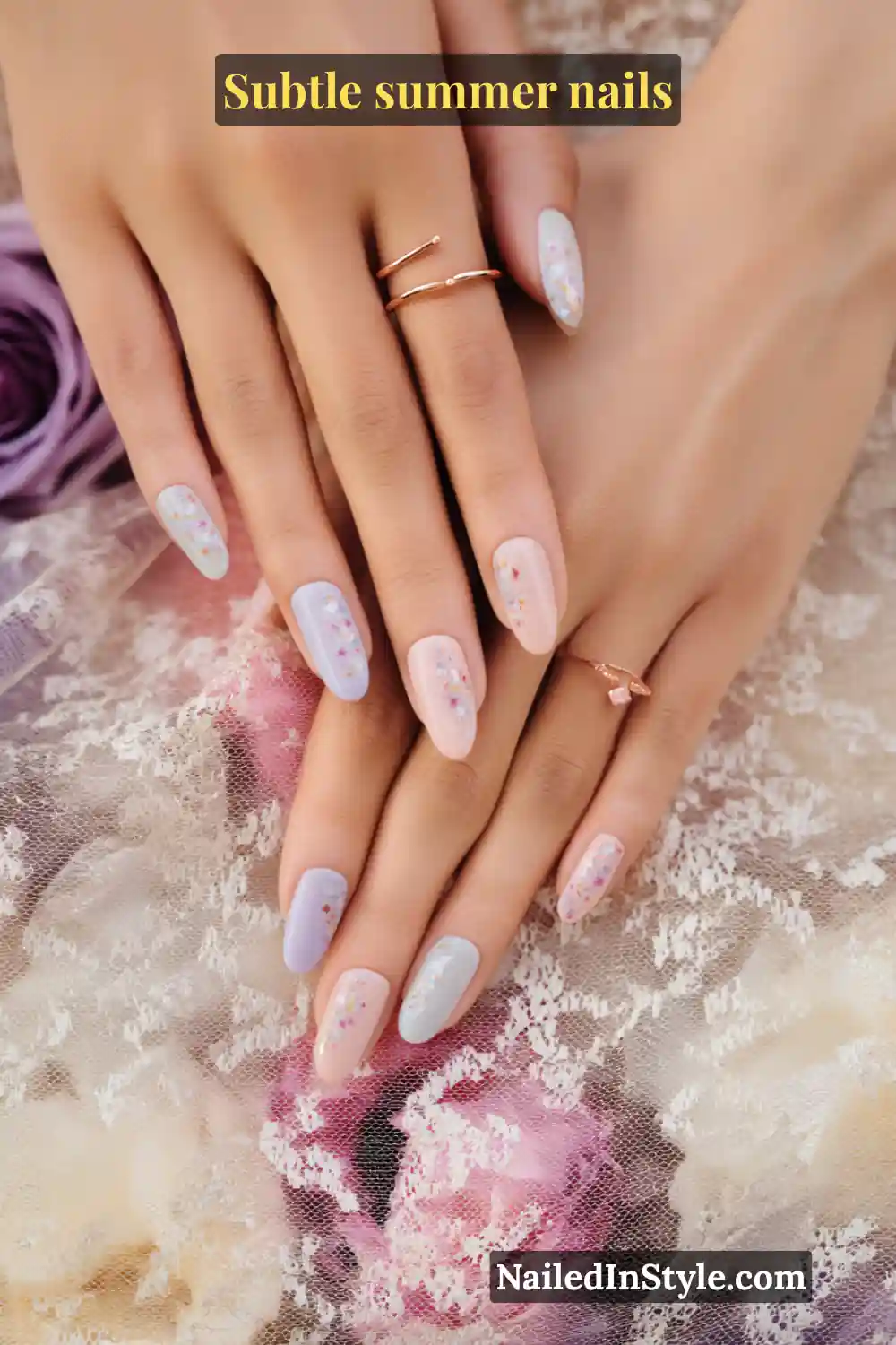 Almond-shaped nails in blush pink, lavender, and lilac with tiny floral confetti accents, styled against lace and roses.