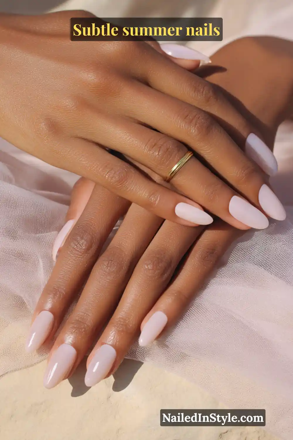 Almond-shaped light pink nails with a glossy finish, styled with a delicate gold band ring, set against airy fabric.
