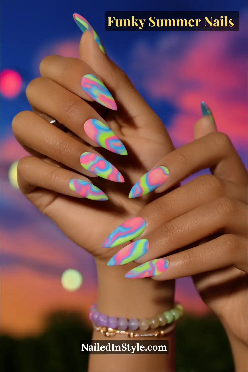 Close-up of long stiletto nails painted in a matte tie-dye swirl design. Neon shades of pink, blue, lime green, and purple blend together in a psychedelic multicolor effect.