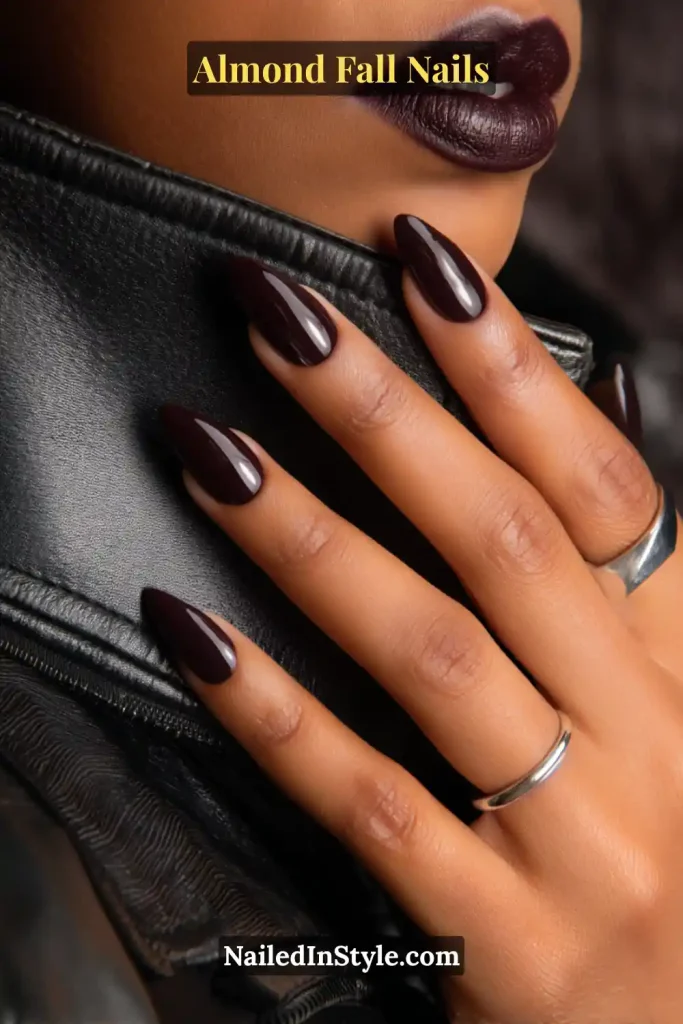 Almond fall nails in deep blackberry purple-black with high-gloss shine, styled with a leather jacket and matching lipstick.