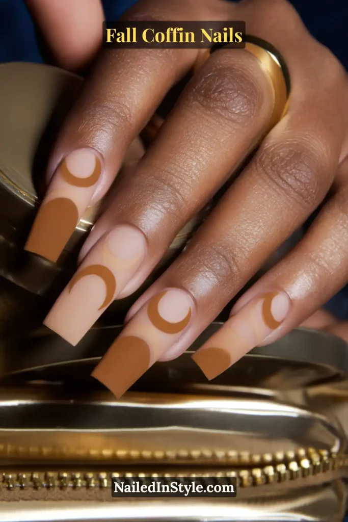 fall coffin nails in caramel half-moon arcs over nude base, bold negative-space style, posed beside a gold zipper detail