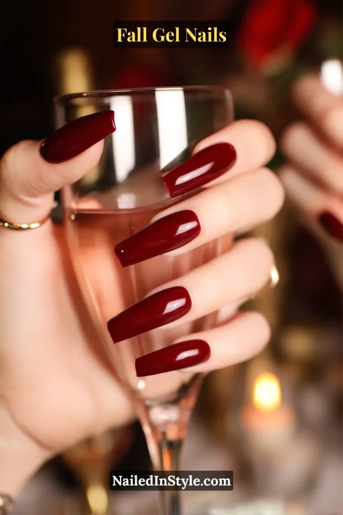 fall gel nails in juicy cranberry wine red, glossy coffin shape holding a dinner glass with warm candlelit background