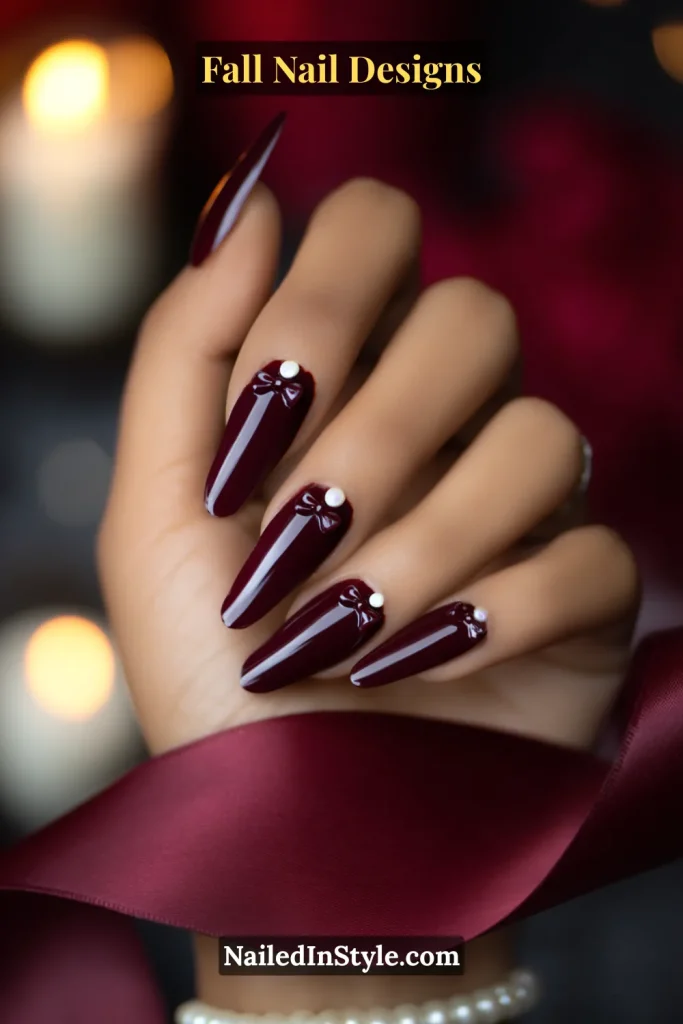 Long almond nails in glossy burgundy with tiny bow decals and pearl studs, hand near burgundy ribbon.