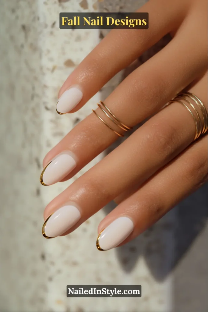 Short oval nails in nude with metallic gold chrome French tips, hand with stacked rings.