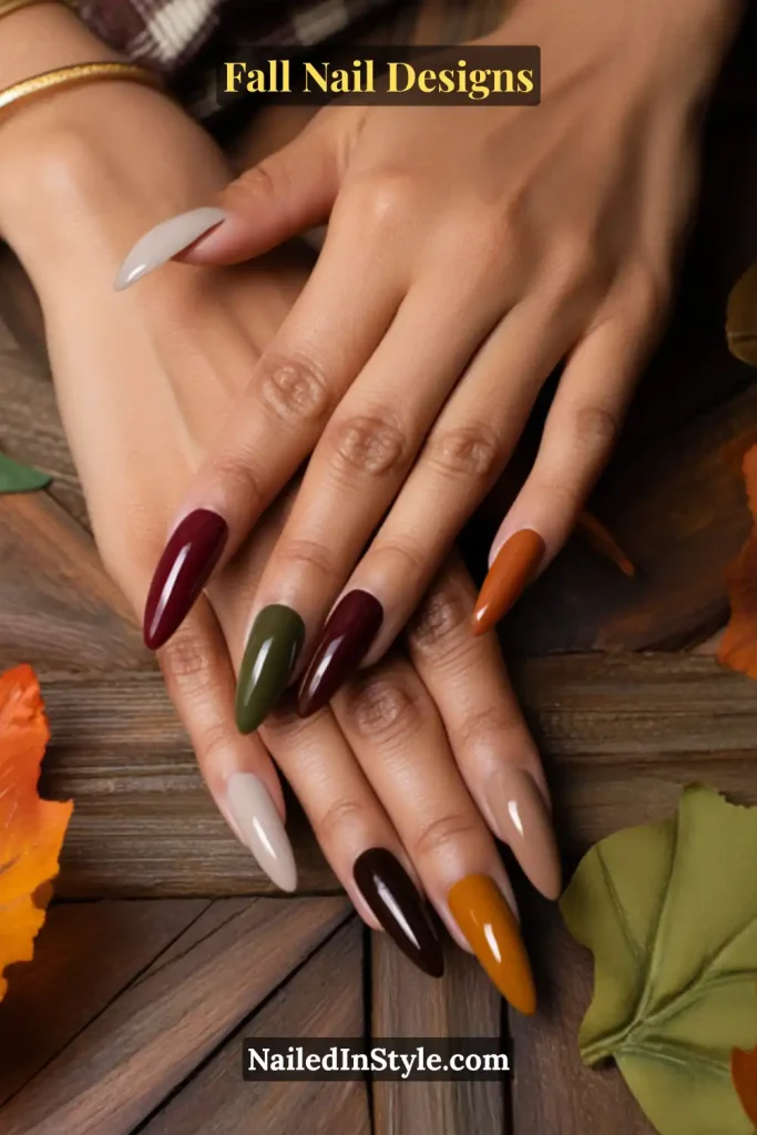 Long almond nails each painted a different fall shade—burgundy, olive, mustard, taupe, and brown—on wood table with leaves.