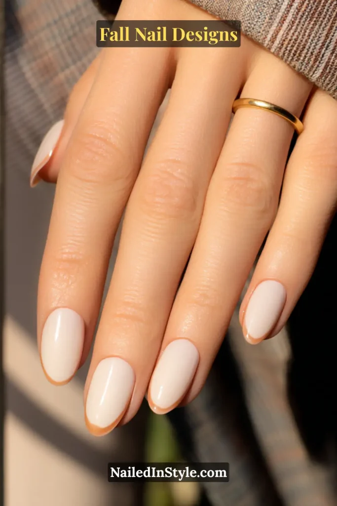 Short oval nails in nude with slim rust French tips, hand on plaid lapel.