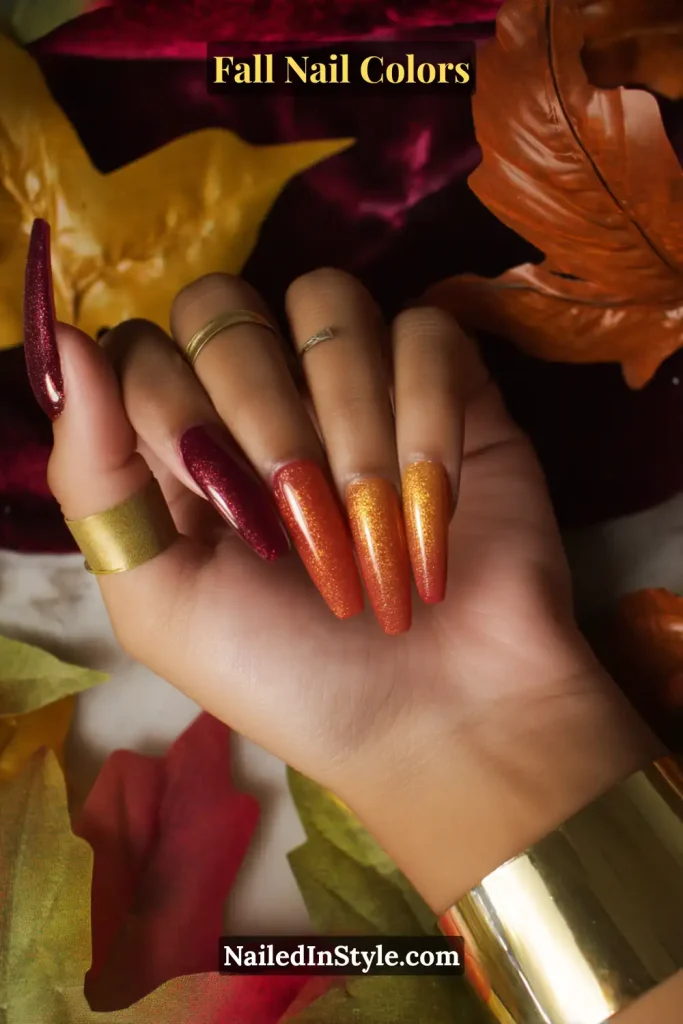 Long ombre nails blending burgundy, pumpkin orange, and gold tones on medium skin, surrounded by autumn leaves.