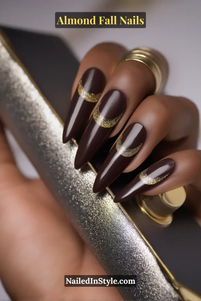 Almond fall nails in deep chocolate brown with fine gold glitter half-moon accents, styled against a metallic clutch.