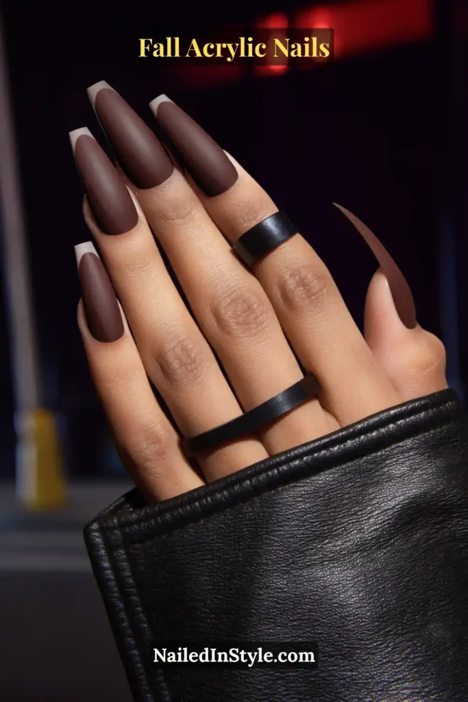 Long square acrylic nails in matte chocolate brown with glossy polished tips, deep skin tone, styled with black rings and leather jacket sleeve at night.