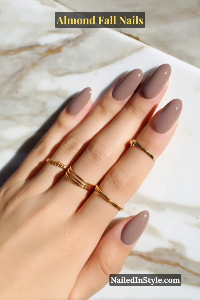 Glossy mauve-taupe almond nails styled with gold rings on a marble surface.