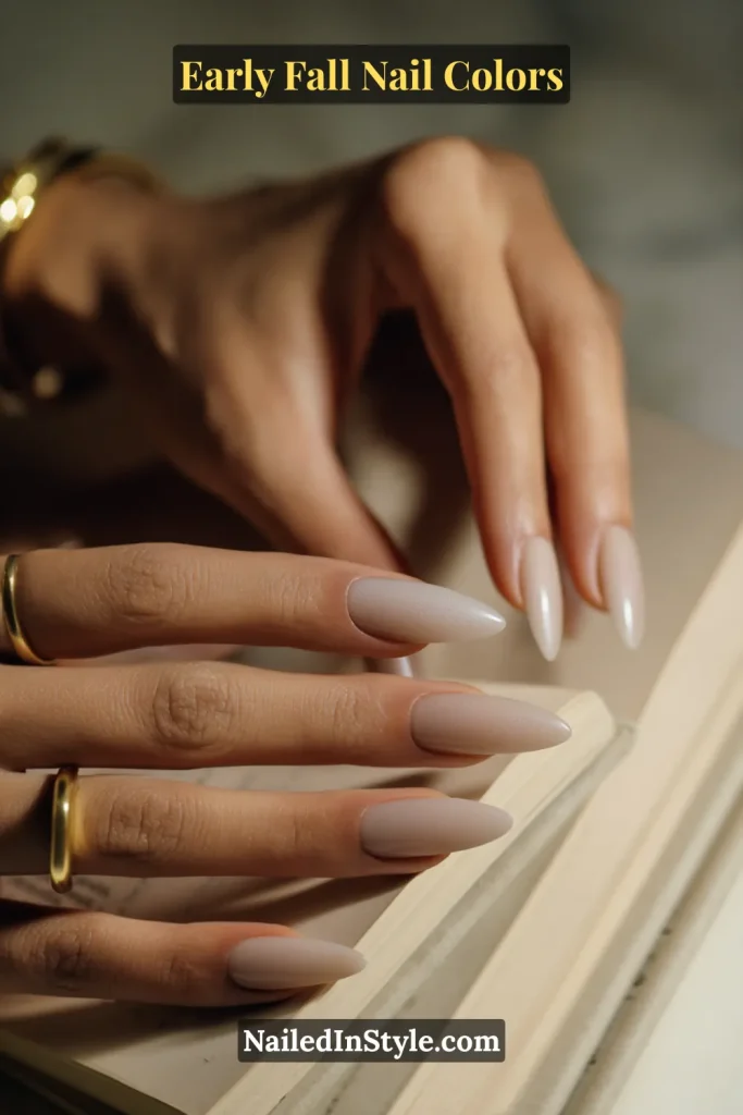 Early fall nail colors in cool mushroom taupe with a soft milky aura glow, styled while turning pages of a linen-bound novel.