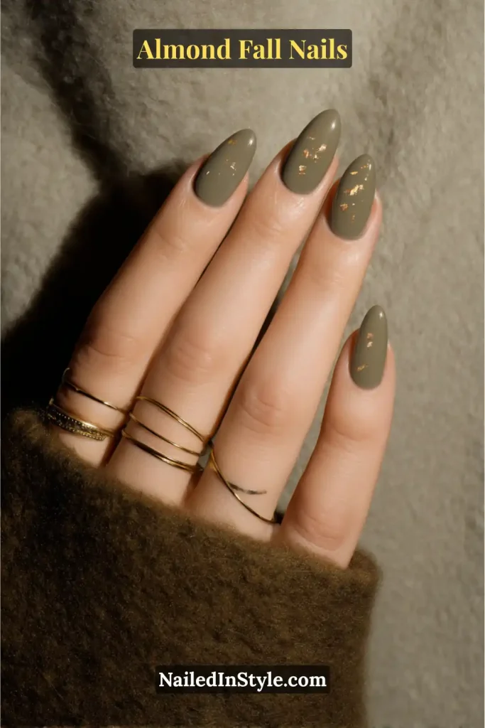 Almond-shaped nails painted in matte olive green with tiny scattered gold flakes, styled with gold rings against a soft wool coat.