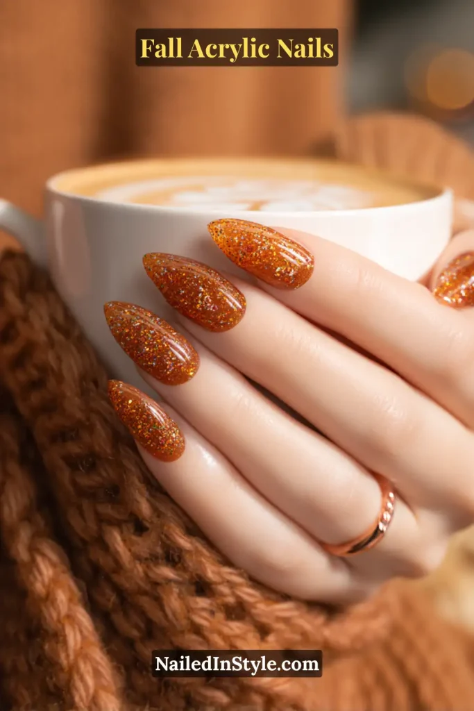 Medium almond acrylic nails with encapsulated pumpkin-orange and gold glitter, shown holding a latte with latte art and cozy knit scarf.