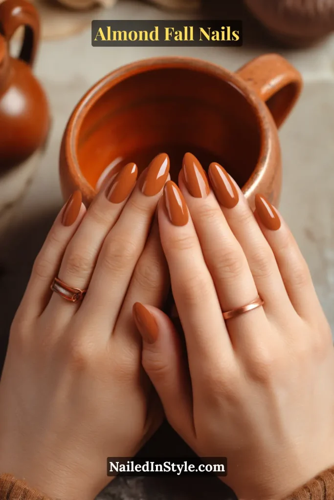 Almond fall nails painted in earthy terracotta orange with a soft glossy finish, styled holding a clay mug.