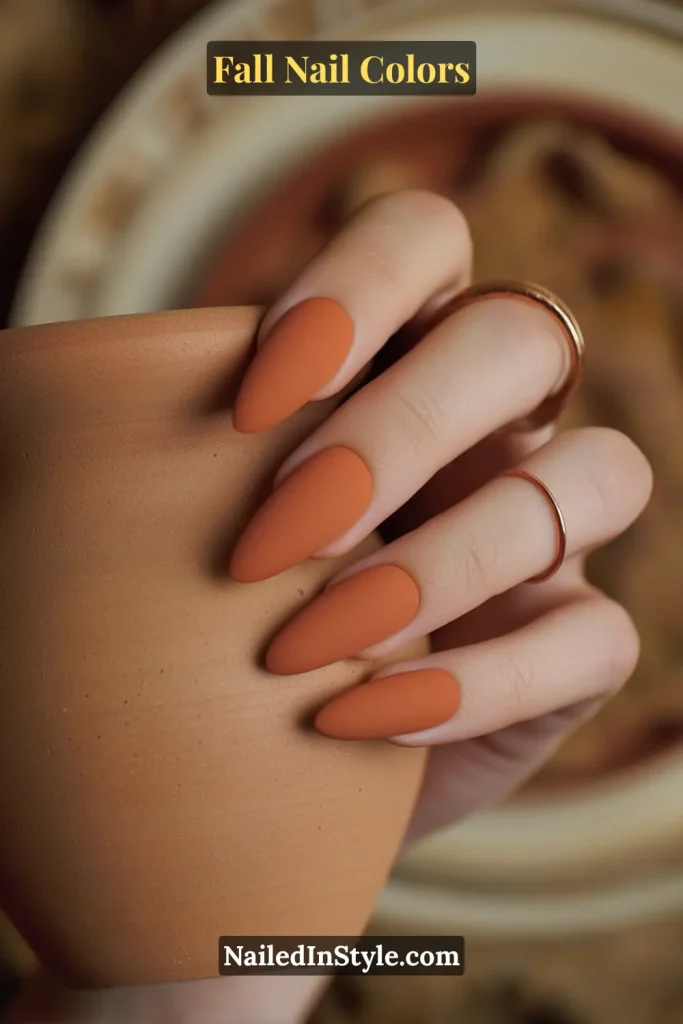 Matte terracotta orange nails on medium skin holding a rustic clay coffee mug, styled with simple gold rings.