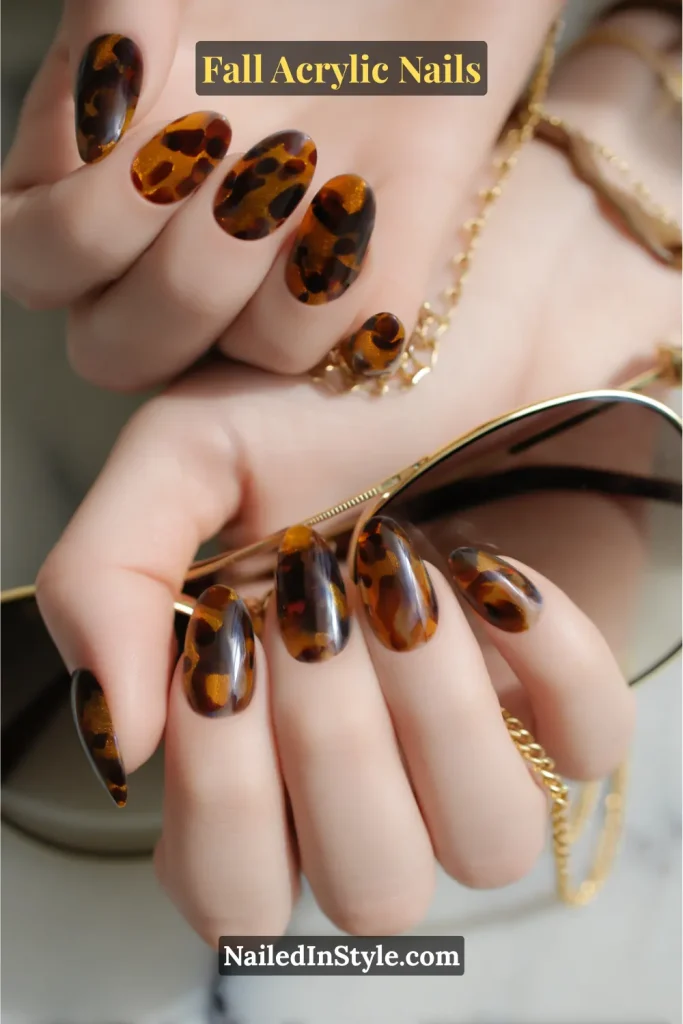 Oval-shaped acrylic nails with tortoiseshell inlay featuring amber, brown, and black patterns on fair to medium skin, styled with gold jewelry and tortoise sunglasses.