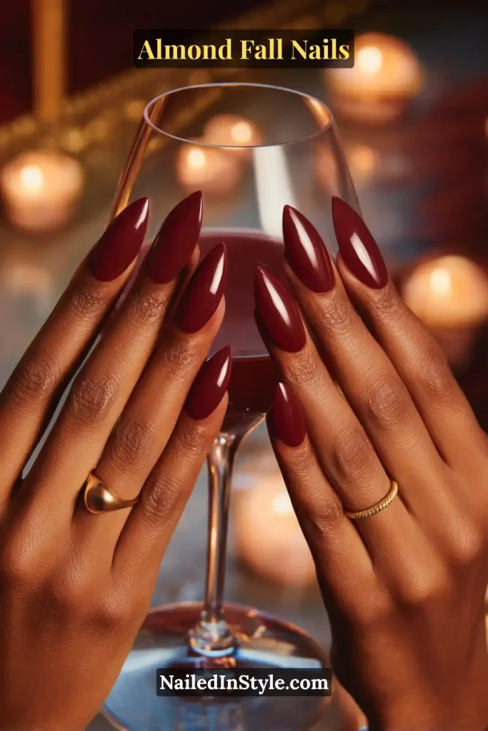 Medium-long almond-shaped nails painted in deep burgundy wine-red polish with a glossy finish, holding a stemmed glass in warm evening light.