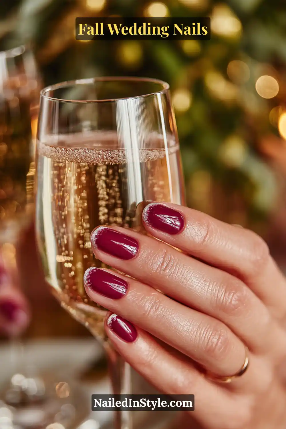 Close-up of fall wedding nails in deep plum polish with natural half-moon cuticle design, hand holding champagne flute.
