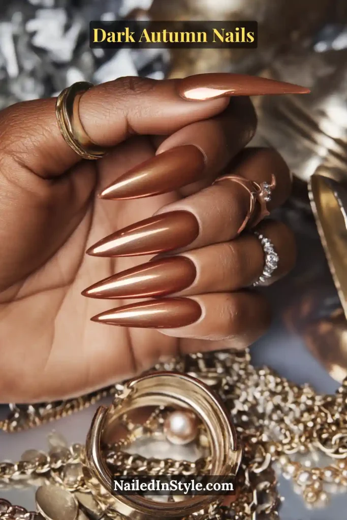 Dark autumn nails in warm bronze chrome with a soft cuticle halo, styled on long stiletto nails against jewelry flatlay with rings and chains.