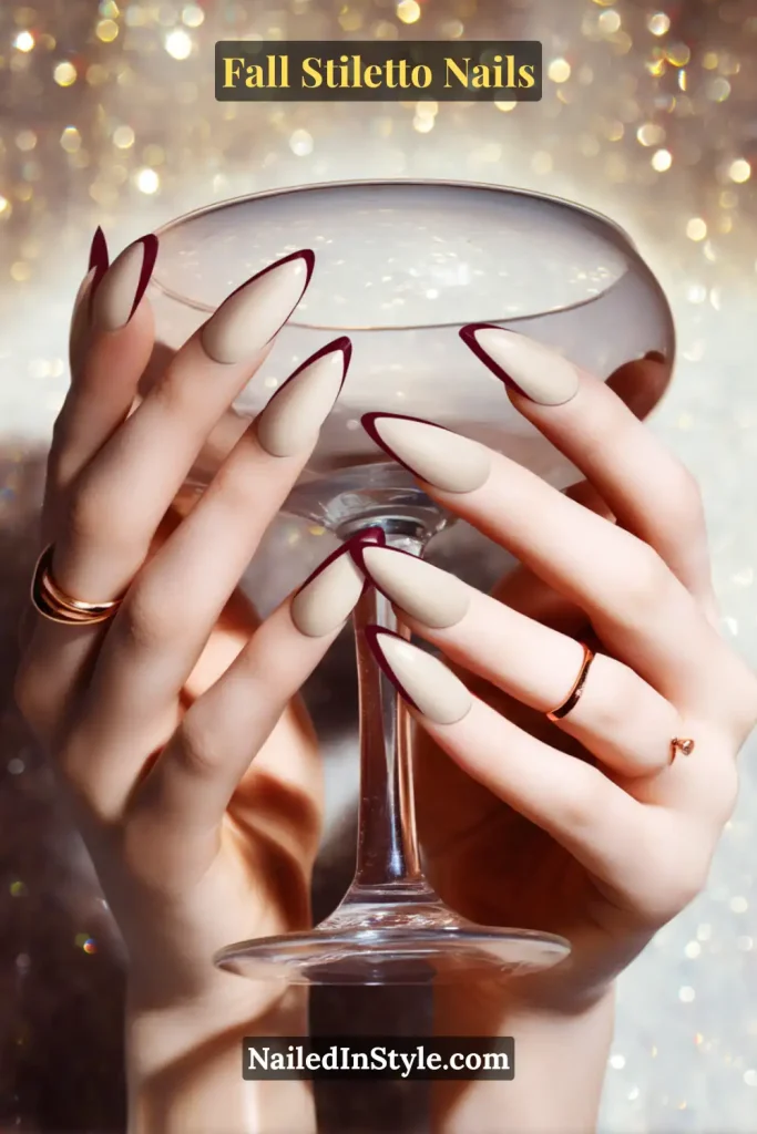 Neutral stiletto nails with slim garnet French tips, styled toasting with a stemmed glass.