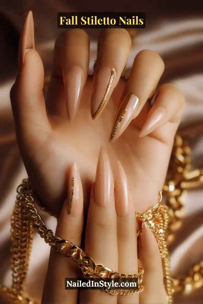 Stiletto nude nails with gold chain accents on select nails, styled with layered gold jewelry.