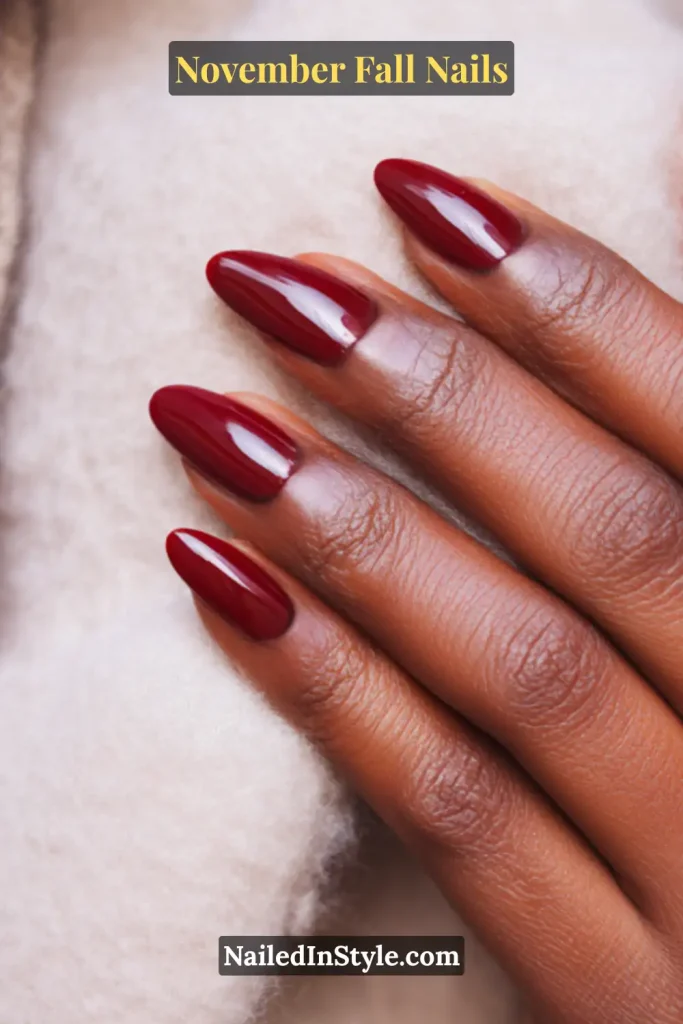Glossy cranberry red November fall nails in a short almond shape, styled against a cozy cashmere sleeve for a warm seasonal vibe.