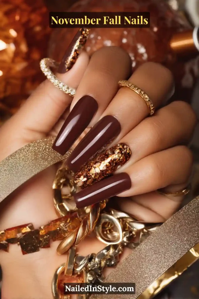 November fall nails in rich espresso brown with a single gold-foil accent nail, styled with layered rings and gold chain jewelry.