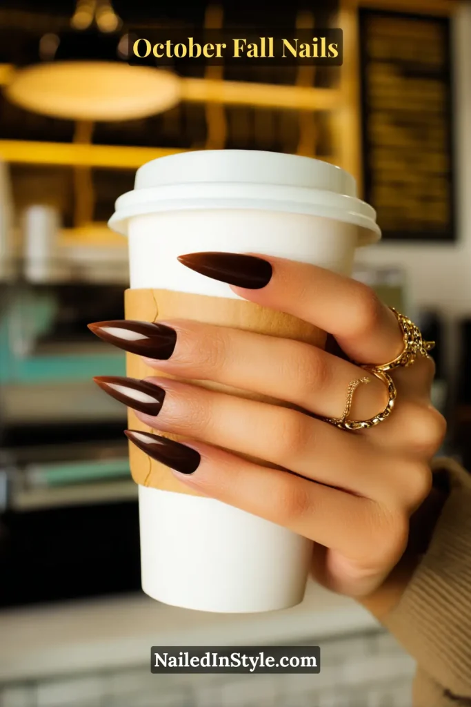 October fall nails in deep espresso coffee crème with a mirror topcoat, styled on stiletto nails holding a latte sleeve with a gold ring accent.