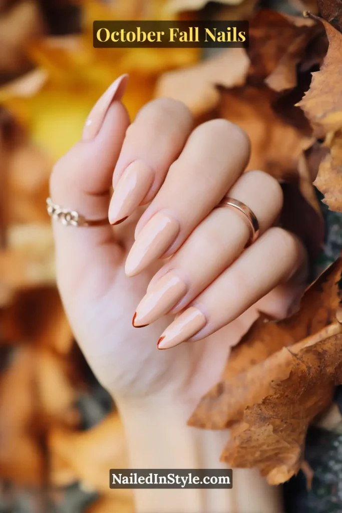 October fall nails in nude polish with tiny maple-colored half-moon accents, almond shape, styled against a backdrop of fallen autumn leaves.
