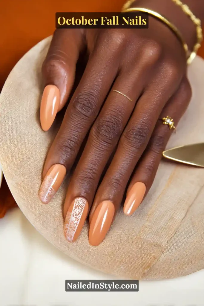 October fall nails in soft pumpkin crème polish with delicate white lace stamping on two accent nails, styled beside a linen napkin and pie server.