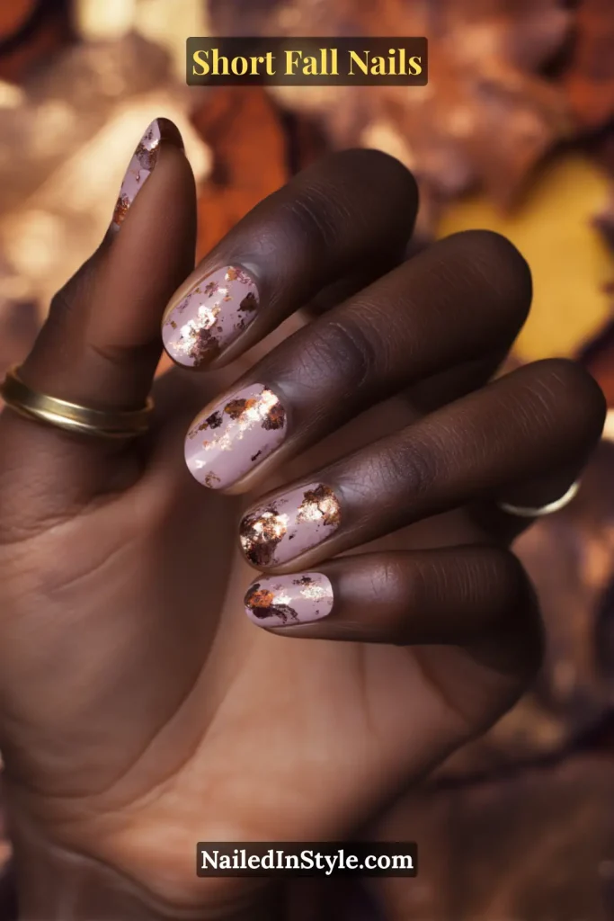 Short neutral nails with scattered bronze foil accents, styled against fallen autumn leaves.