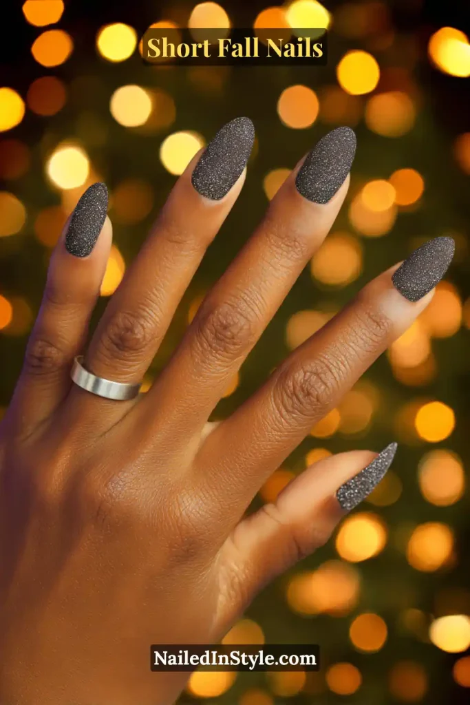 Short almond nails in charcoal jelly with superfine glitter, glowing softly against twinkle light bokeh.