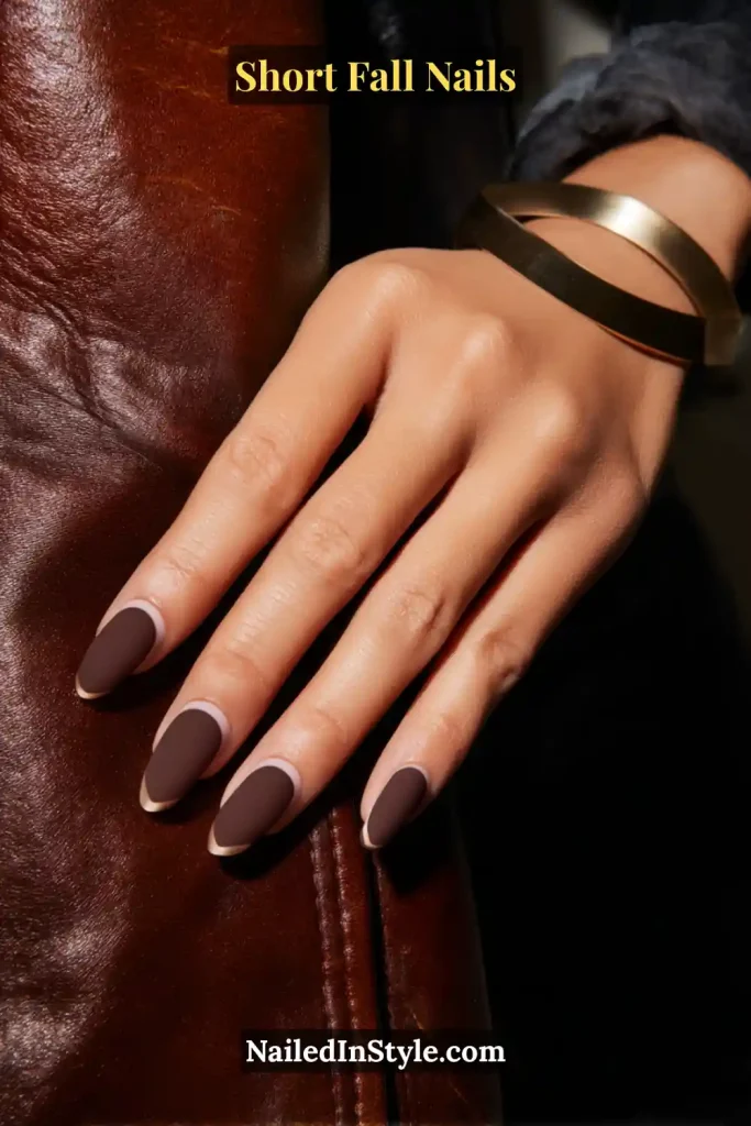 Short almond nails in suede-matte mocha with a glossy outline, styled against a leather jacket sleeve.