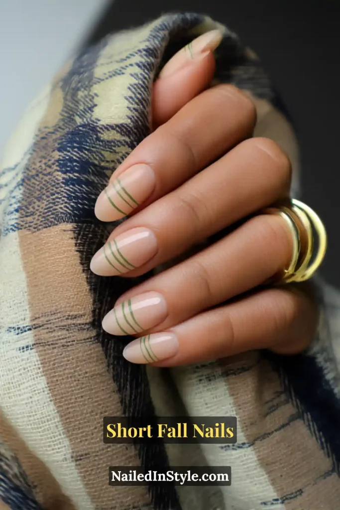Short fall nails with a sheer nude base and slim olive French tips, styled against a cozy plaid scarf.