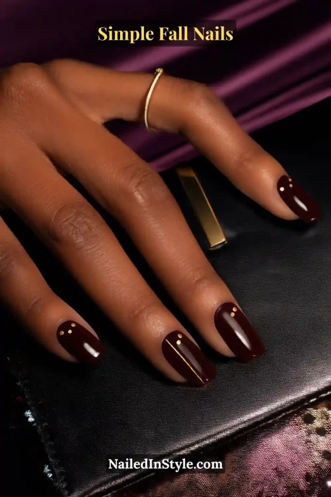 Simple fall nails in a deep black-cherry polish with micro gold dots on each nail and a vertical gold line accent on the ring finger, styled with a satin clutch clasp.