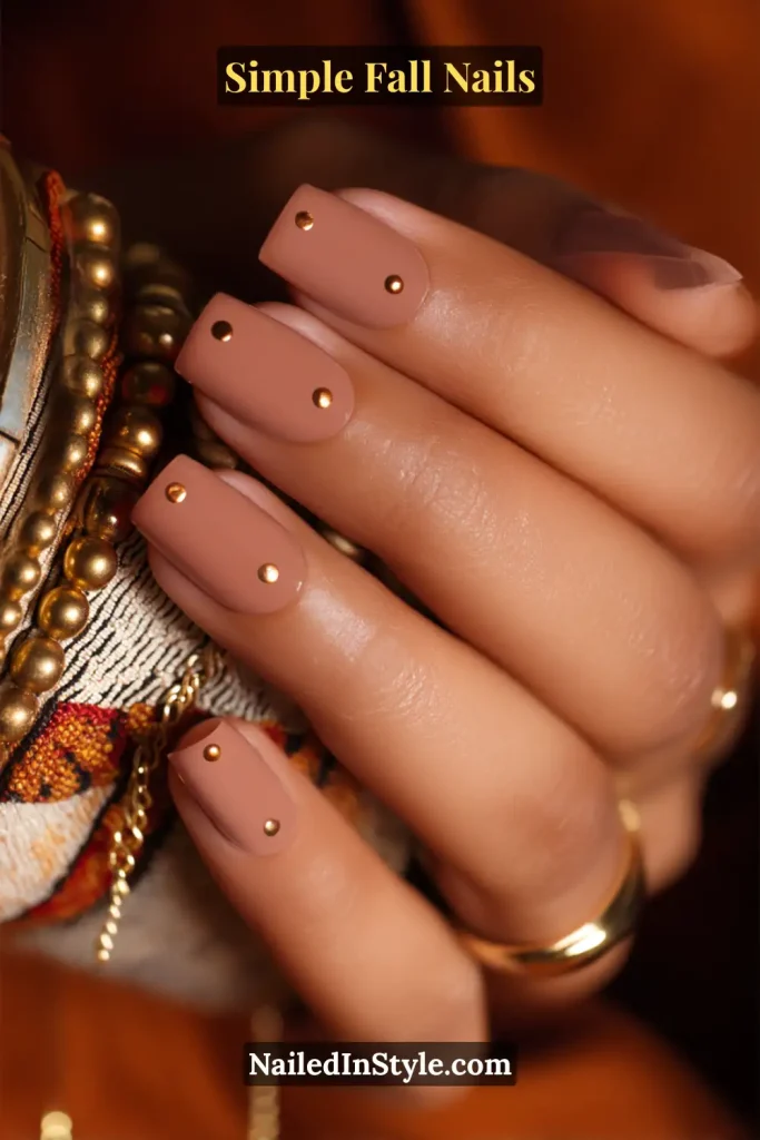 Simple fall nails with a nude base and a single bronze dot near the cuticle, styled in a jewelry flatlay with rings and chain accents.